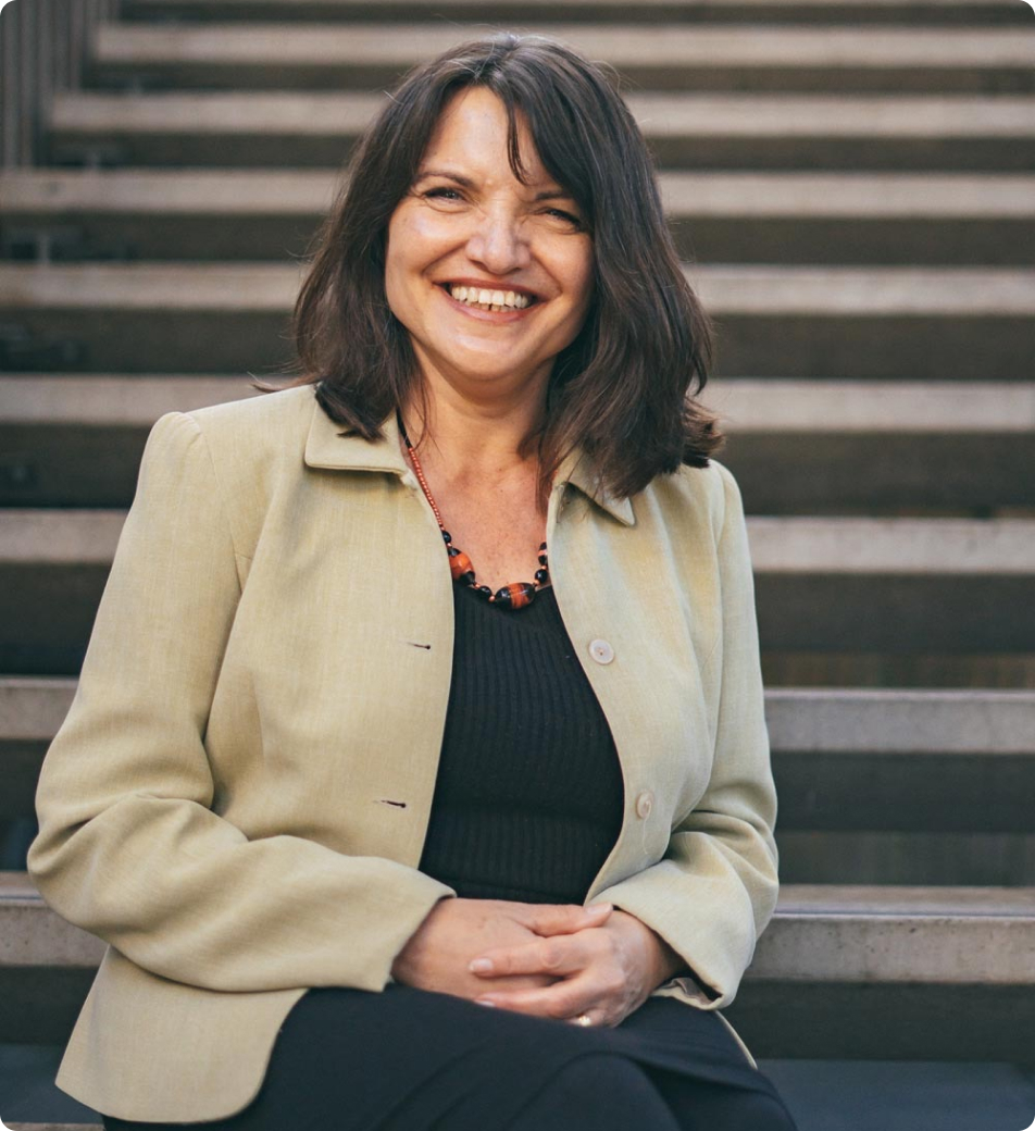 Image depicts a woman with a genuine smile, seated on steps. She's dressed in a light olive blazer, black top, and trousers, accented with a statement necklace and brown boots, blending professionalism with personal style against an outdoor, architectural backdrop.