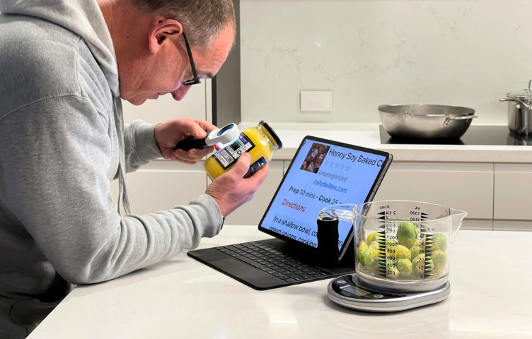 A man leans across a marble kitchen counter, holding a magnifier to read the label on a jar. A set of talking measuring scales sits next to his laptop, with a recipe zoomed in on the screen.