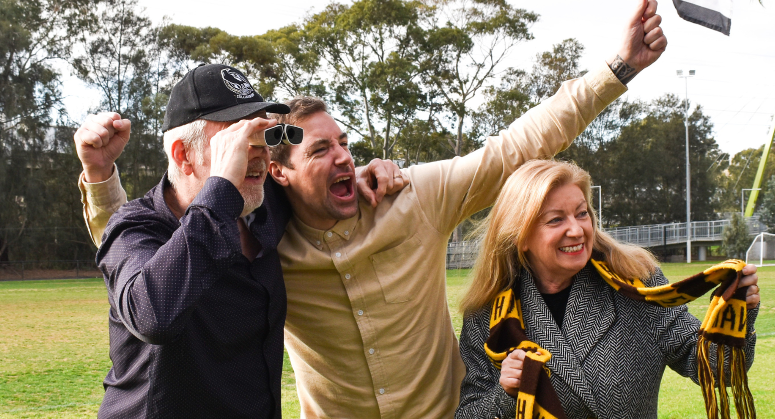 Three people stand cheering behind the rails at a local football game, dressed in their AFL team colours. One man wearing an AFL hat is watching the game through a pair of binoculars.