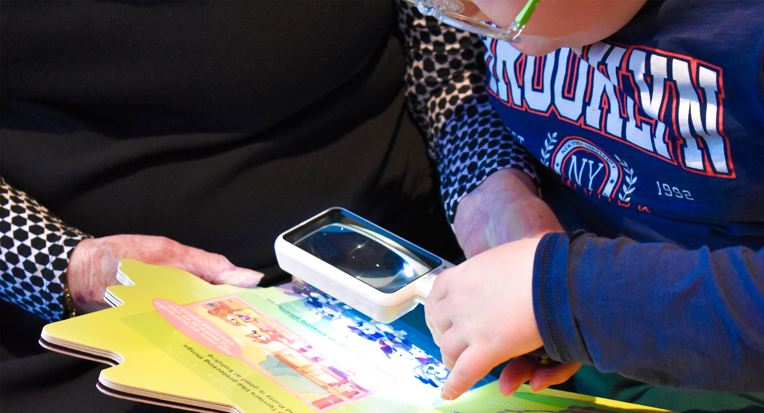 A young boy and his grandmother sit together to read a children’s book together. They both hold an LED magnifier over the words to read the book easily.