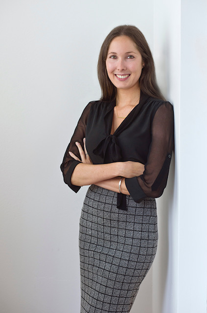 "Caroline leaning against a white wall in business attire."