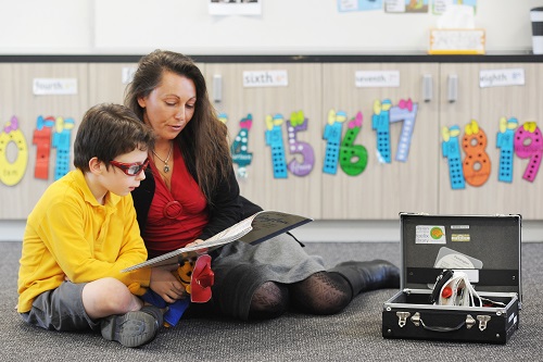 A young boy sits on the floor reading a large print book with his teacher