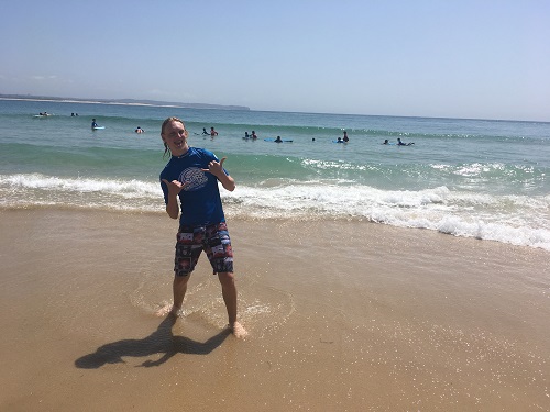 A young male Vision Australia client stands on the sand just before the waterline looking at the camera