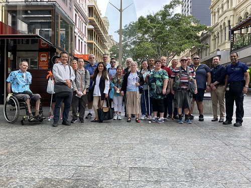 A group of people, some holding white canes and other mobility aids stand in a pedestrian mall