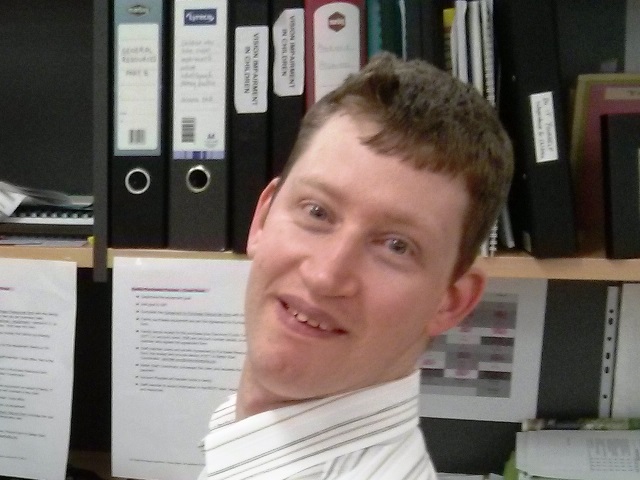 Volunteer Paul McIntosh sitting in front of his computer at work with files on a shelf in the background