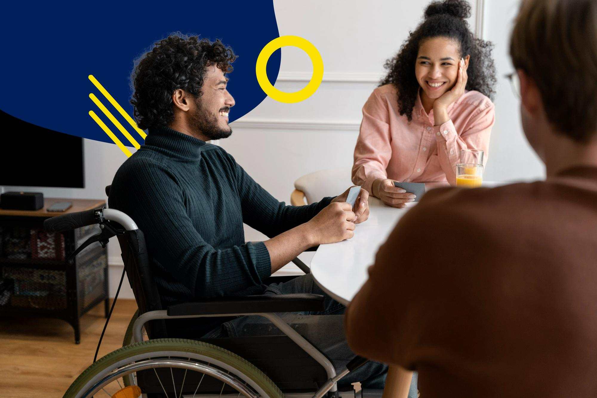 Three friends sharing a laugh while playing cards at home, one of whom is in a wheelchair.