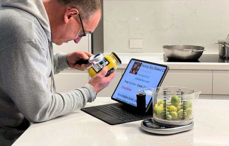 A man leans across a marble kitchen counter, holding a magnifier to read the label on a jar. A set of talking measuring scales sits next to his laptop, with a recipe zoomed in on the screen.