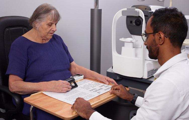 A lady in a purple t-shirt sits with her eye doctor in his office, using a magnifier to read a sheet of paper. 