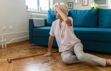 A woman in a pink top sits awkwardly on the floor with her hand on her forehead, glasses and cane fallen beside her. 