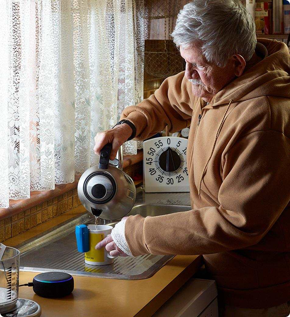 An older man in a brown hoodie pours hot water from a kettle into a cup at the kitchen sink. A talking clock and a smart speaker are nearby.