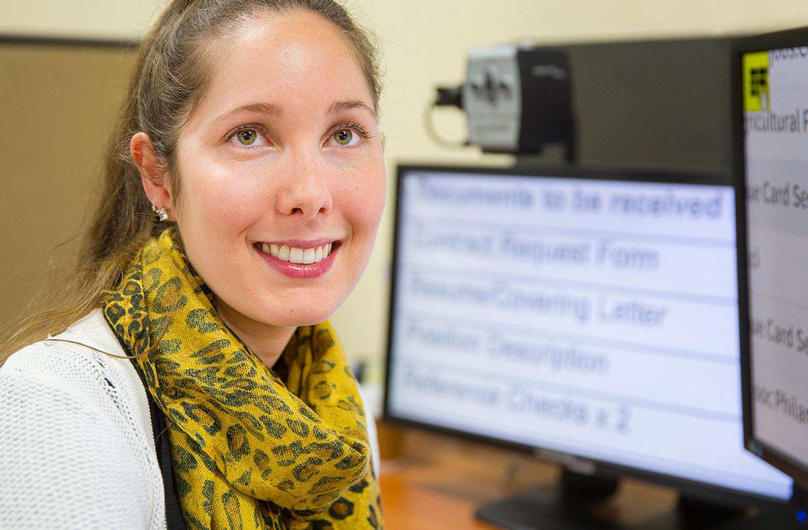 A smiling young woman sits at a computer with enlarged text on the monitor.