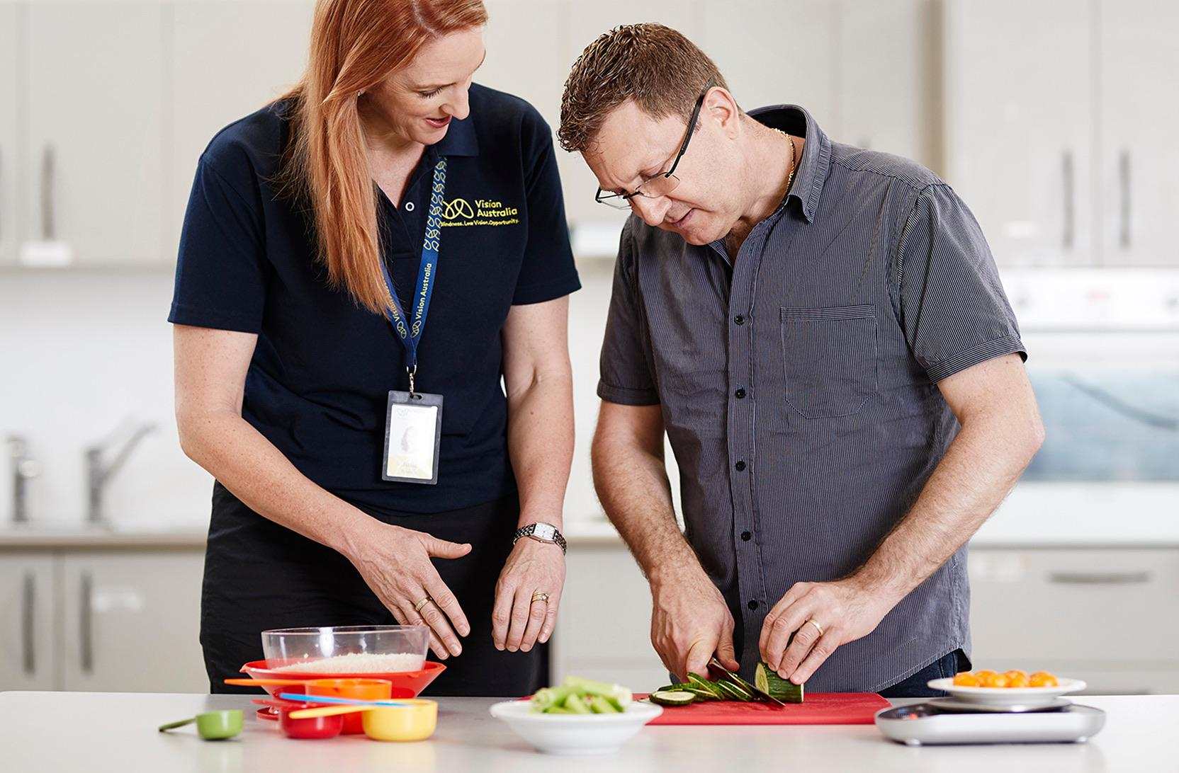 A woman in a Vision Australia polo shirt assists a man slicing cucumbers in a kitchen with measuring cups and ingredients.