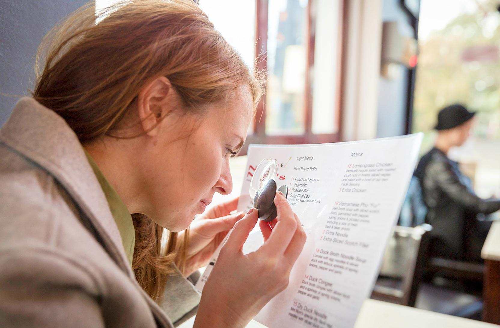 A young woman holds a magnifier close to a restaurant menu, focusing on the text.