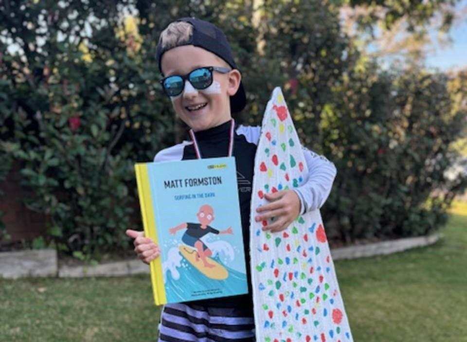 Hunter is wearing sunglasses and a hat, and has zinc cream on his nose. He is holding a book and a surfboard.