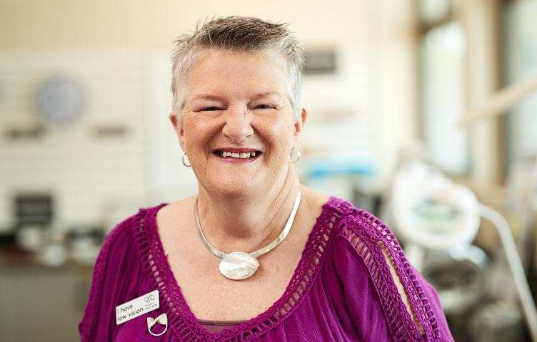 A lady in a purple top and shell necklace is smiling to camera, wearing a ‘I have low vision’ badge. 