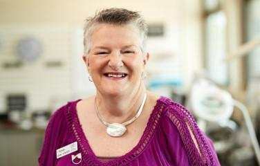 A smiling woman with short grey hair, wearing a purple T-shirt, an ‘I Have Low Vision’ badge, and a striking shell necklace, looks at the camera.