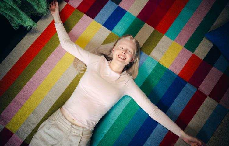 A blonde woman lies on her back on a colourful rug, arms spread wide and smiling to camera.
