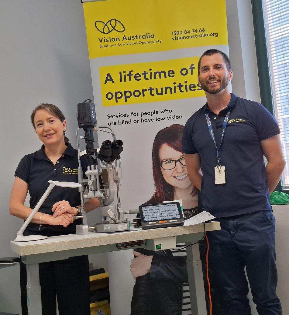 A Vision Australia optometrist and occupational therapist are smiling and standing behind a desk with assistive technology at the Know Your Vision clinic at the Sydney Eye Hospital.