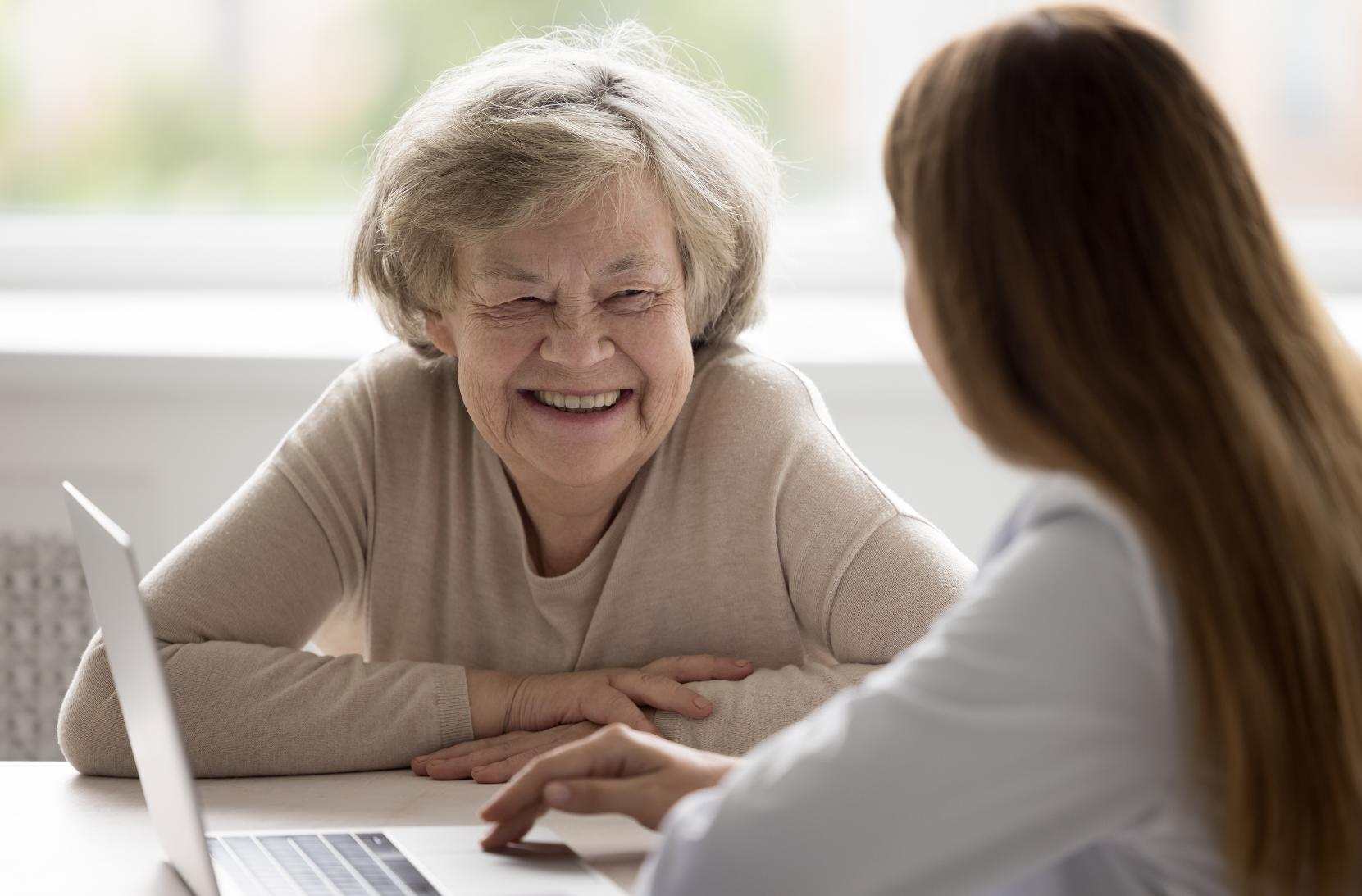 An older lady in a beige top smiles at her female support worker, sitting in front of an open laptop. 