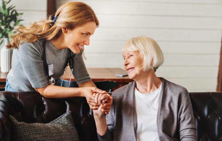 An older lady in a grey cardigan is holding the hand of her female doctor, in conversation and smiling. 